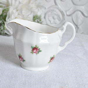 Może przedstawiać: Biały ceramiczny dzbanek do śmietanki ze złotą obwódką i kwiatowym wzorem różowych róż. Dzbanek ma uchwyt i zaokrąglone ciało.