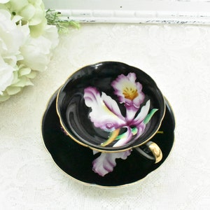 May include: Black teacup and saucer set with a gold rim and handle. The cup and saucer feature a painted white and purple orchid design. The set is placed on a white lace tablecloth, with white flowers in the background.
