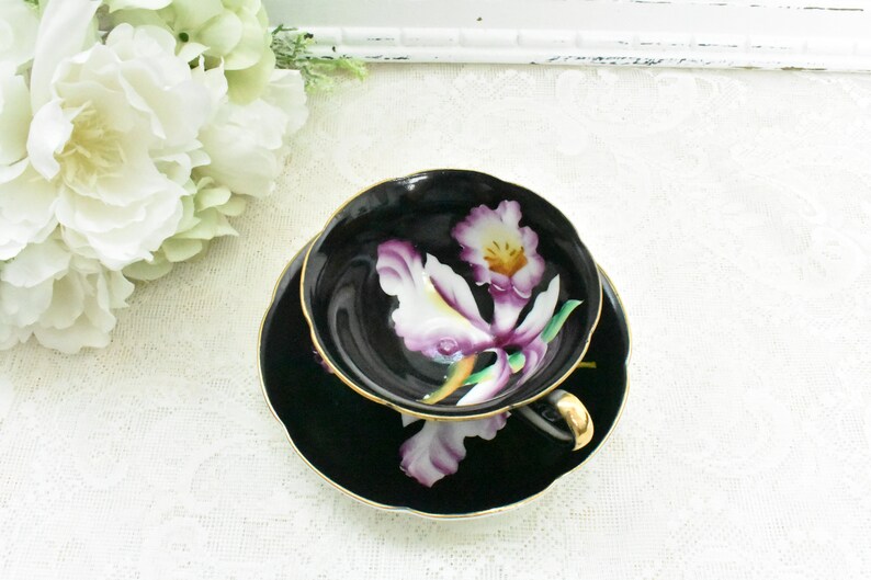 May include: Black teacup and saucer set with a gold rim and handle. The cup and saucer feature a painted white and purple orchid design. The set is placed on a white lace tablecloth, with white flowers in the background.