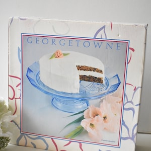 Puede incluir: Una caja blanca con un plato para pastel azul y blanco con un pastel glaseado. La caja tiene el texto "GEORGETOWNE" y las palabras "Footed Cakeplate" en la parte inferior.