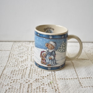 May include: Cream-colored ceramic mug with a winter scene featuring two teddy bears in a snowy landscape. The mug has a blue border with snowflakes and the word "Believe" inside the rim. A beige handle completes the design.