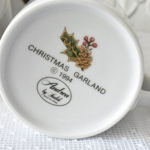May include: White ceramic mug with a Christmas Garland design. The mug features a green holly sprig with red berries and a yellow flower. The text "Christmas Garland &copy; 1994" is printed on the mug. The bottom of the mug has the text "Andrea by Sadek Made in Japan."