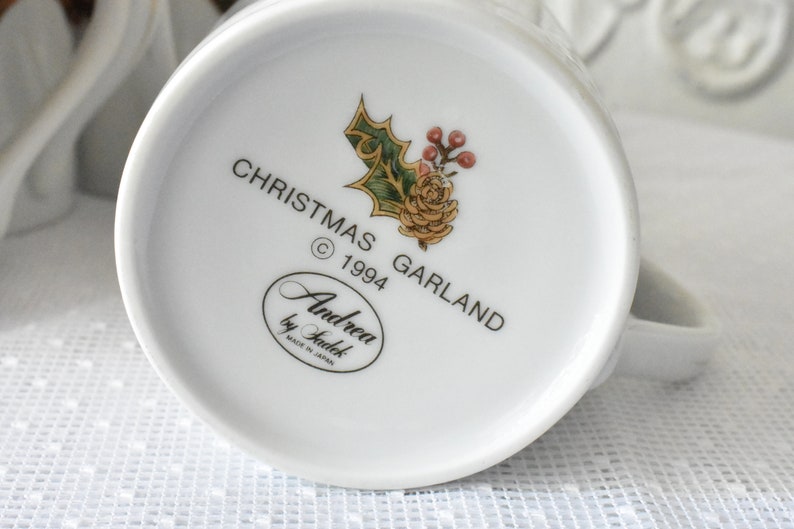 May include: White ceramic mug with a Christmas Garland design. The mug features a green holly sprig with red berries and a yellow flower. The text "Christmas Garland &copy; 1994" is printed on the mug. The bottom of the mug has the text "Andrea by Sadek Made in Japan."