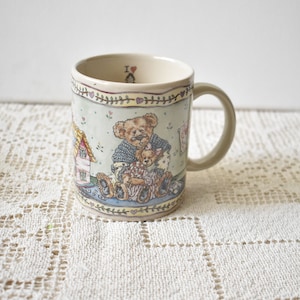 May include: A ceramic mug with a light green background and a whimsical illustration of teddy bears. The mug features a beige handle and rim, with a decorative border of hearts and leaves. The words "I ❤️" are visible inside the rim.