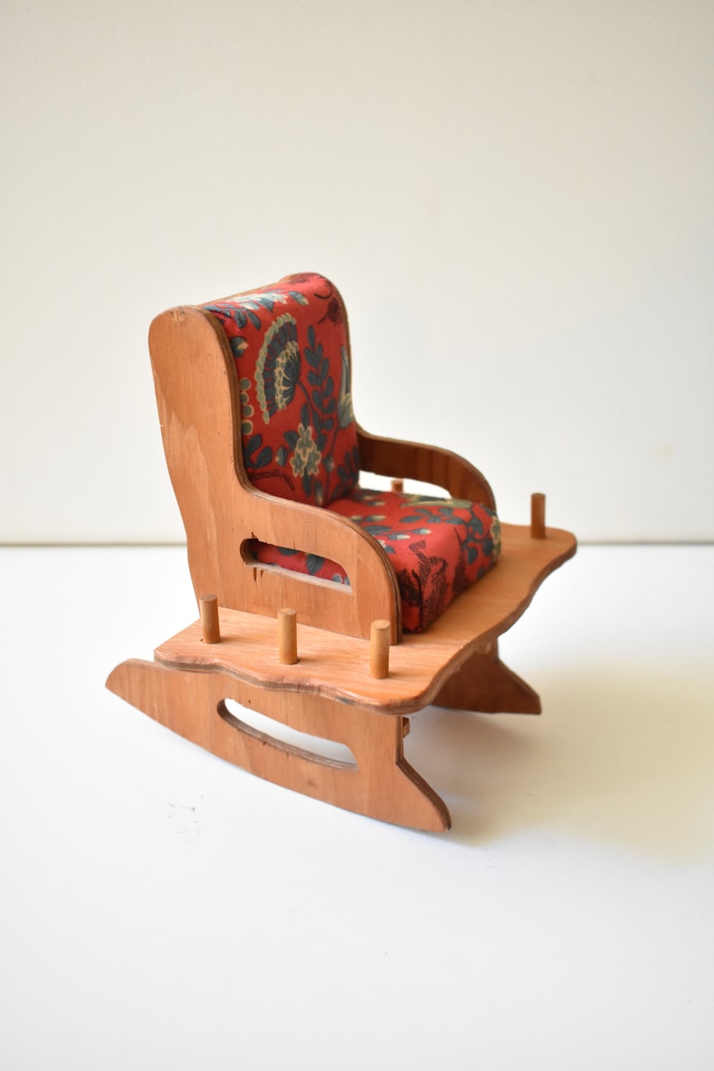 May include: A wooden rocking chair with a red floral patterned cushion. The chair is made of light brown wood and has a simple design.