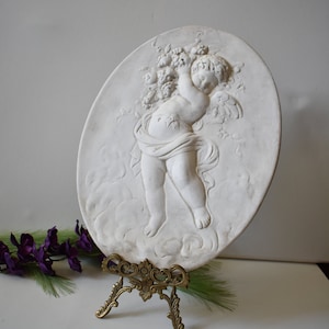 Puede incluir: Placa de pared de cerámica blanca con un querubín sosteniendo un racimo de uvas. La placa es de forma ovalada y tiene un diseño en relieve tridimensional. Se muestra en un soporte de metal dorado.