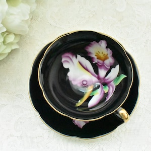 May include: A black teacup and saucer set with a gold trim. The teacup and saucer feature a painted orchid design in shades of purple, white, and yellow. The teacup has a gold handle. The set is on a white lace tablecloth.