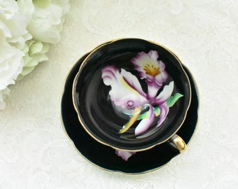 Puede incluir: Un juego de taza y platillo negro con ribete dorado. La taza y el platillo presentan un diseño de orquídea pintada en tonos morado, blanco y amarillo. La taza tiene un asa dorada. El juego está sobre un mantel de encaje blanco.