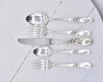 May include: Five pieces of silver flatware, including a fork, knife, and three spoons, are arranged in a row on a white surface. Each piece has an ornate floral design.