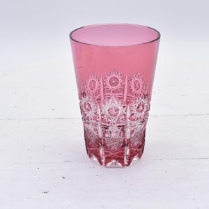 May include: A tall, clear glass with a pink hue and intricate cut-glass design. The glass has a rounded base and a straight, cylindrical body.