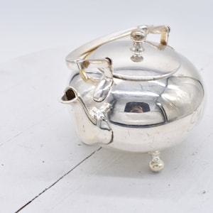 May include: A silver teapot with a gold handle and a round body. The teapot has a lid and a spout. It is sitting on a white surface.