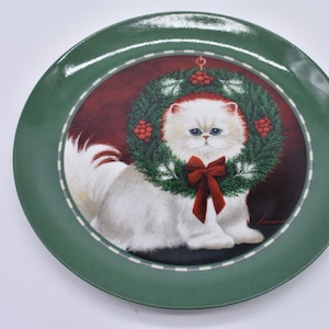 May include: A green-rimmed decorative plate featuring a white Persian cat wearing a Christmas wreath with a red bow. The cat has blue eyes and is centered on a burgundy background.