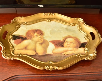 May include: A gold-colored tray with a raised edge and two handles. The tray features a painting of two cherubs with wings, one looking thoughtful and the other looking to the side.