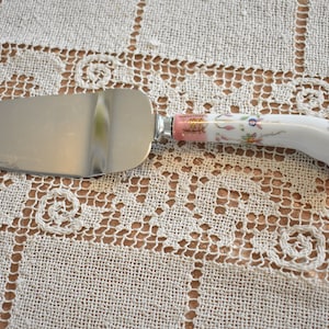 Puede incluir: Un servidor de pastel de plata con un mango de porcelana blanca decorado con diseños florales rosas y dorados.