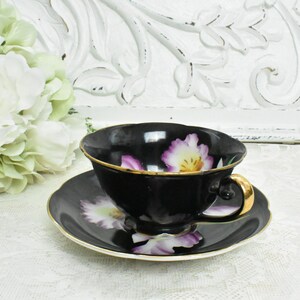 May include: A black teacup and saucer set with a gold rim and handle. The cup and saucer feature a floral design with purple and yellow flowers. The set is placed on a white lace tablecloth.