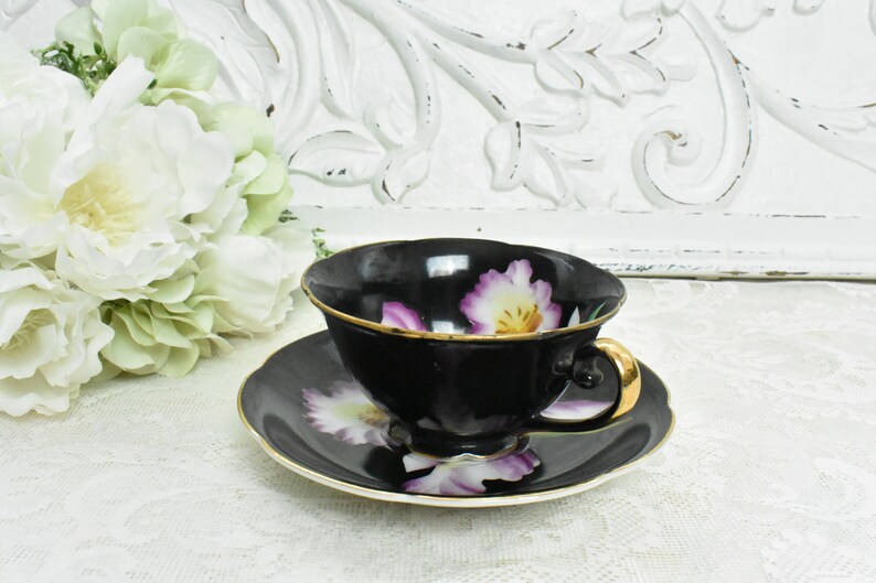 May include: A black teacup and saucer set with a gold rim and handle. The cup and saucer feature a floral design with purple and yellow flowers. The set is placed on a white lace tablecloth.