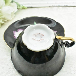 May include: Black teacup and saucer set with a gold handle and trim. The saucer and the outside of the cup are black, while the inside of the cup is white. The saucer has a floral design with purple and green accents.