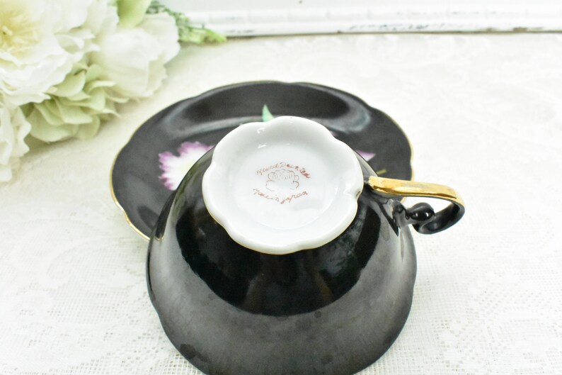 May include: Black teacup and saucer set with a gold handle and trim. The saucer and the outside of the cup are black, while the inside of the cup is white. The saucer has a floral design with purple and green accents.