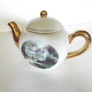 May include: White porcelain teapot with a gold handle and lid. The teapot features a painting of a house with the text "Home is Where The Heart Is" on the lid.