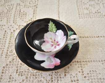 Puede incluir: Un juego de taza y platillo negro con un diseño floral rosa y blanco. La taza y el platillo tienen un borde dorado.
