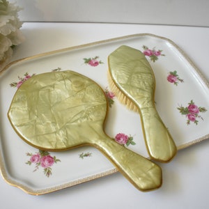 May include: A vintage vanity set with a green, pearlescent hand mirror and a matching brush. The set is on a white tray with a floral pattern.
