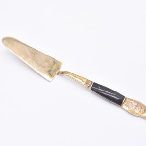 May include: A gold-toned cake server with a black handle. The handle has a decorative design.