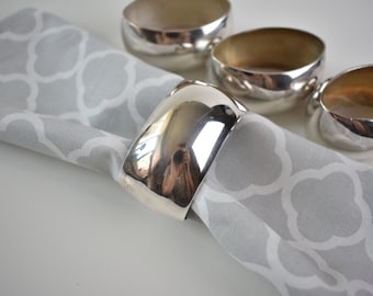 May include: Four silver napkin rings, one of which is on a gray patterned napkin. The rings are smooth and shiny, and they have a rounded shape.