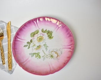 May include: A pink and white porcelain plate with a floral design. The plate has a scalloped edge and is decorated with white roses and green leaves.