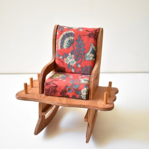 May include: A miniature rocking chair made of wood, featuring a floral patterned cushion and a small table with wooden dowels. The chair is a scaled-down version of a traditional rocking chair.