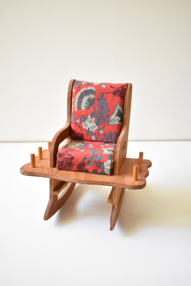 May include: A miniature rocking chair made of wood, featuring a floral patterned cushion and a small table with wooden dowels. The chair is a scaled-down version of a traditional rocking chair.