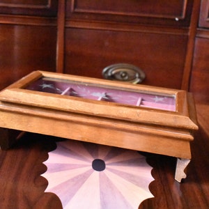 May include: A small, wooden jewelry box with a glass top, resting on a decorative table. The box has a light brown finish and a pink interior. The table has a patterned design in shades of pink and brown.