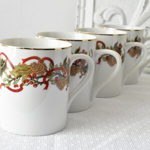 May include: Set of four white ceramic mugs with gold trim and a festive holiday design featuring red ribbons, green holly leaves, and brown pine cones.