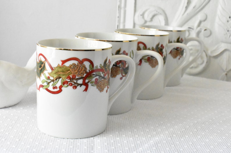 May include: Set of four white ceramic mugs with gold trim and a festive holiday design featuring red ribbons, green holly leaves, and brown pine cones.