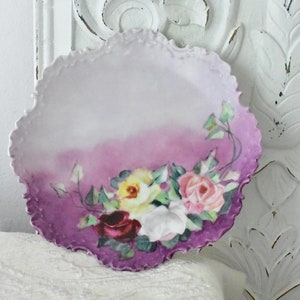 May include: A vintage porcelain plate with a scalloped edge and a hand-painted design of pink, yellow, and red roses on a purple background.