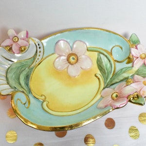 May include: A decorative ceramic dish with a light blue, yellow and green floral design. The dish has a raised rim and is decorated with gold accents.