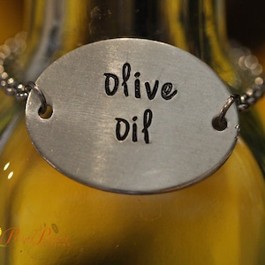 May include: A brushed silver oval metal tag with the words olive oil stamped in black ink, suspended by a silver chain around the neck of a glass bottle containing a yellow liquid. The tag features two small circular holes for the chain attachment.