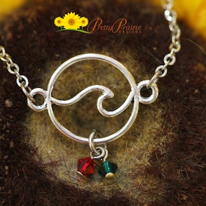May include: Silver necklace with a circle pendant featuring a wave design. The pendant has two small dangling crystals, one red and one green.