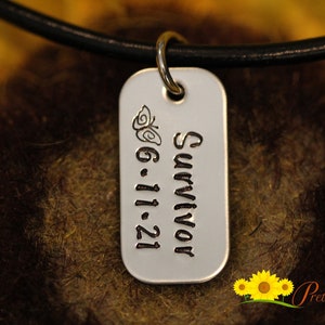 May include: A silver dog tag pendant with the word "Survivor" and the date "6.11.21" engraved in black. A butterfly design is above the date. The tag hangs from a black leather cord. The background is a blurred mix of brown and yellow.