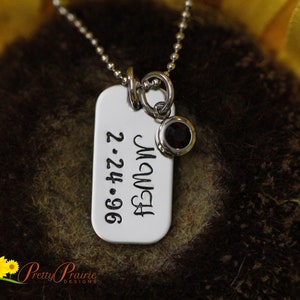 May include: A silver-toned rectangular dog tag pendant on a ball chain necklace. The pendant is engraved with the text "MAWMA" and the date "2.24.96". A small circular charm with a dark red gemstone is attached to the same jump ring as the tag.