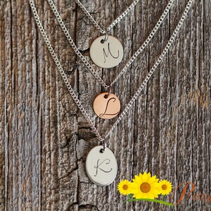 May include: Three silver chains with round pendants, each engraved with a different letter: K, L, and M. The chains are hanging against a wood background.