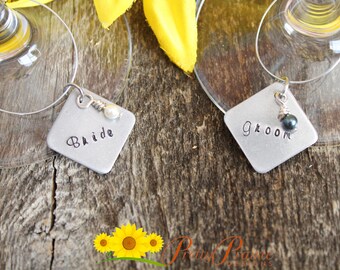 May include: Two silver metal wine glass charms with square tags. The tag on the left says "Bride" and has a white pearl. The tag on the right says "Groom" and has a black pearl.
