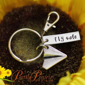 Pode incluir: Chaveiro de prata com um amuleto de avi&atilde;o de papel e uma etiqueta que diz "Fly safe". O chaveiro est&aacute; em um fundo de girassol.