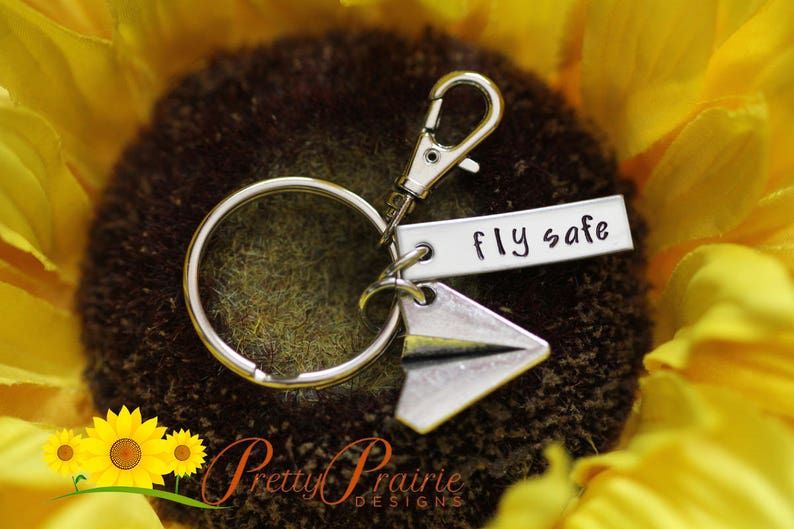 Pode incluir: Chaveiro de prata com um amuleto de avi&atilde;o de papel e uma etiqueta que diz "Fly safe". O chaveiro est&aacute; em um fundo de girassol.