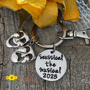 May include: A silver-toned keychain featuring a round charm with the text "Seussical the Musical 2025". The keychain also includes two theater mask charms and a lobster clasp.