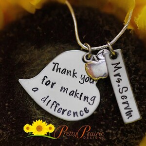 May include: Silver necklace with a heart charm that says "Thank you for making a difference" and a tag charm that says "Mrs. Servin". The charms are attached to a silver chain.