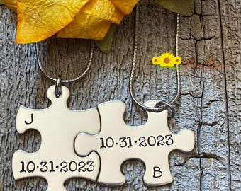 May include: Two silver-toned metal puzzle piece pendants on thin snake chains. The left piece is stamped with the letter J and the date 10.31.2023. The right piece is stamped with the date 10.31.2023 and the letter B. The pieces interlock together.