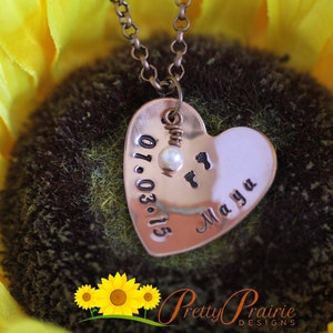 May include: A copper heart-shaped pendant necklace with a pearl and the date 01.03.15 engraved on one side and the name Maya on the other side. The necklace is photographed on a sunflower.