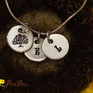 May include: A silver necklace with three charms. The charms are round and have a tree, the letter E, and the letter L engraved on them. The necklace is on a brown background.