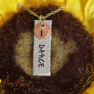 May include: A silver necklace with a hammered metal pendant that says "Dance" and a copper disc with the letter "I" on it. The necklace is photographed against a yellow sunflower.