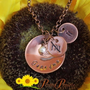 May include: Copper necklace with three charms. The largest charm is round and has the words "Grandma" engraved on it. The middle charm is round and has the Roman numeral "XII" engraved on it. The smallest charm is round and has the number "II" engraved on it. A small pearl bead is attached to the charm with the words "Grandma".